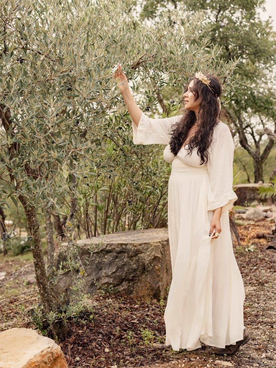 Camila Mello in an olive grove, wearing cream, reaching up to a branch