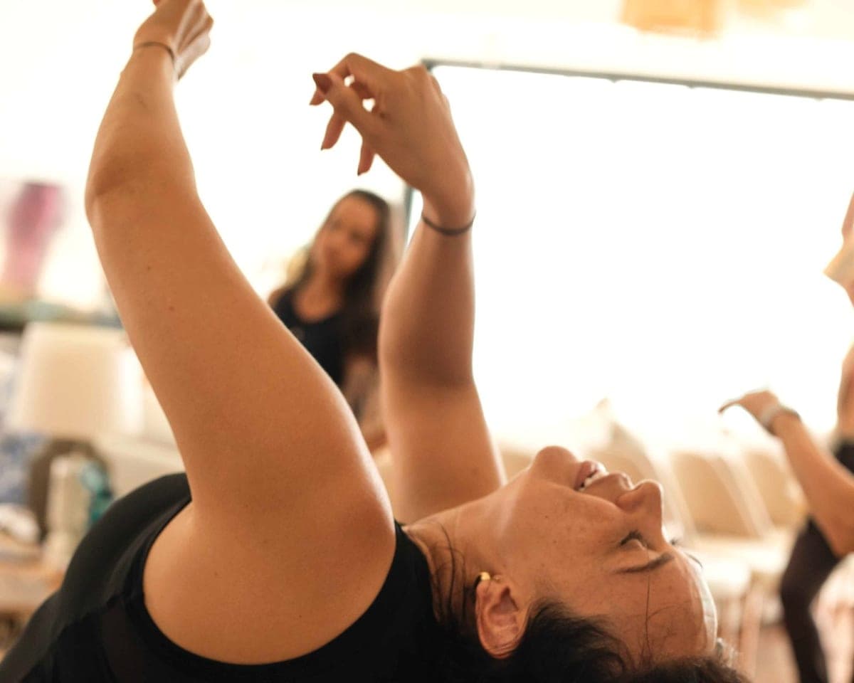 Camila Mello arching back with both arms raised, in bright window light