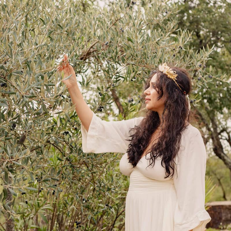 Camila Mello in a cream dress reaching up to an olive branch