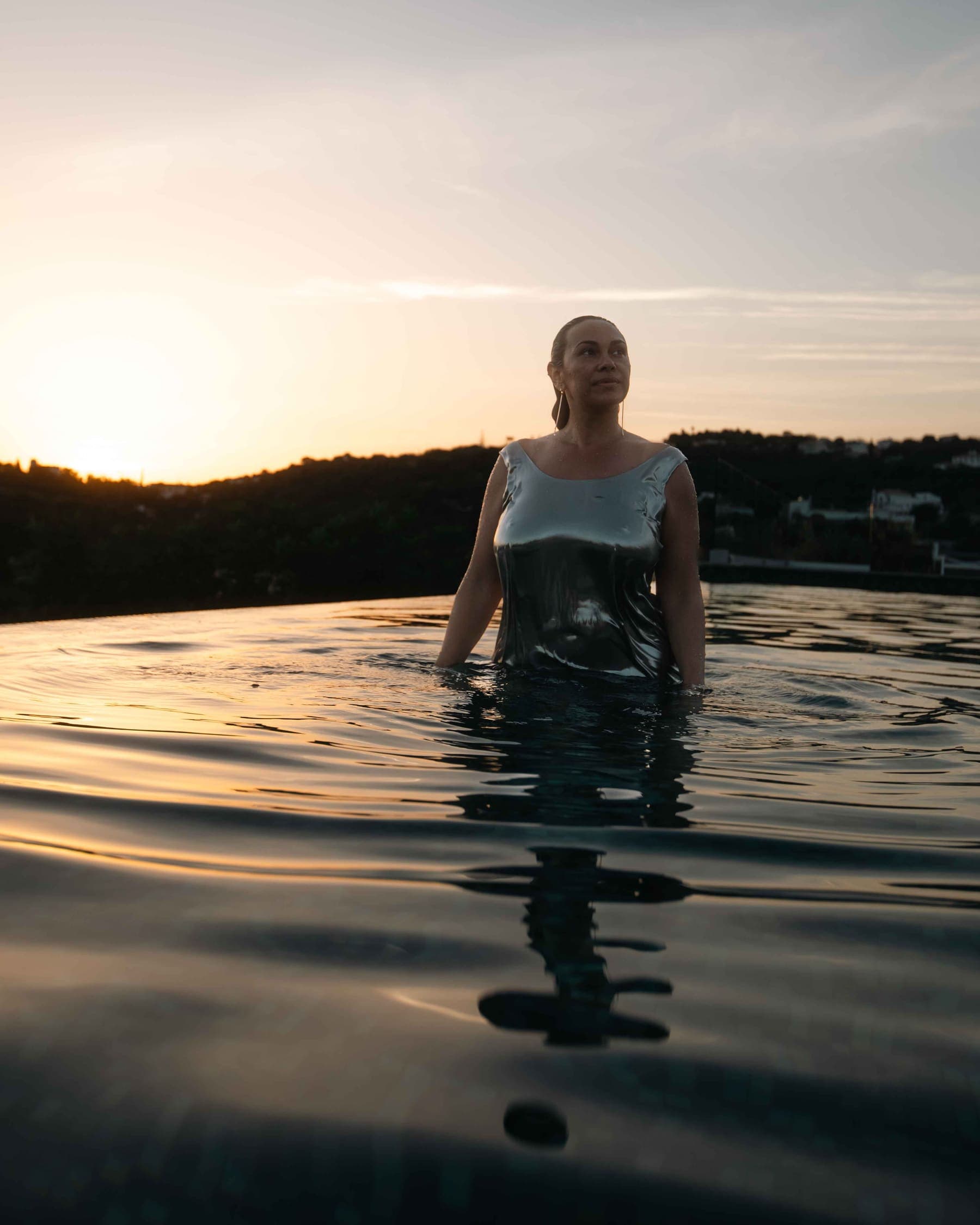 Camila Mello standing waist-deep in an infinity pool at sunset, in a silver dress