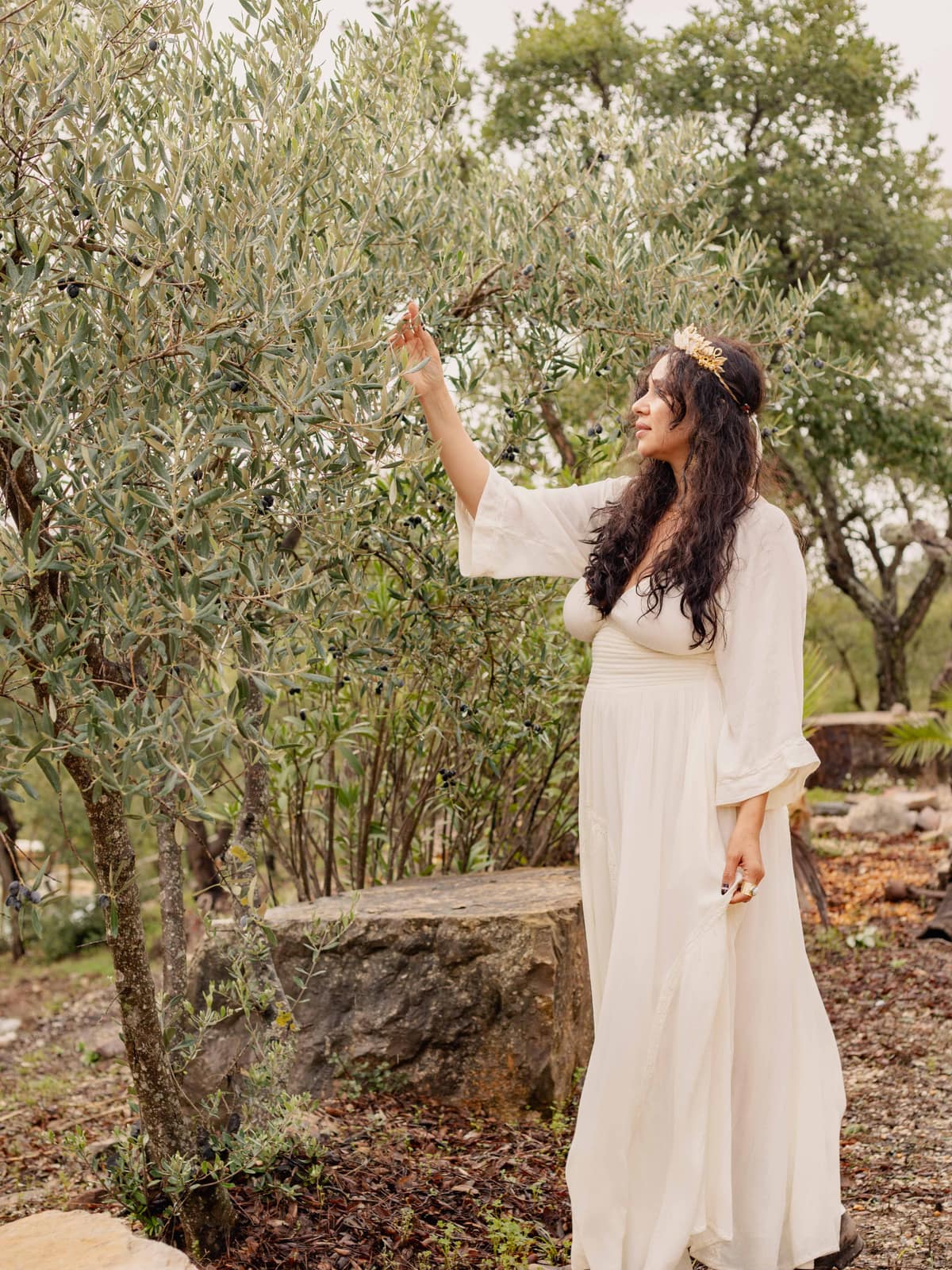 Camila Mello in an olive grove, reaching up to a branch
