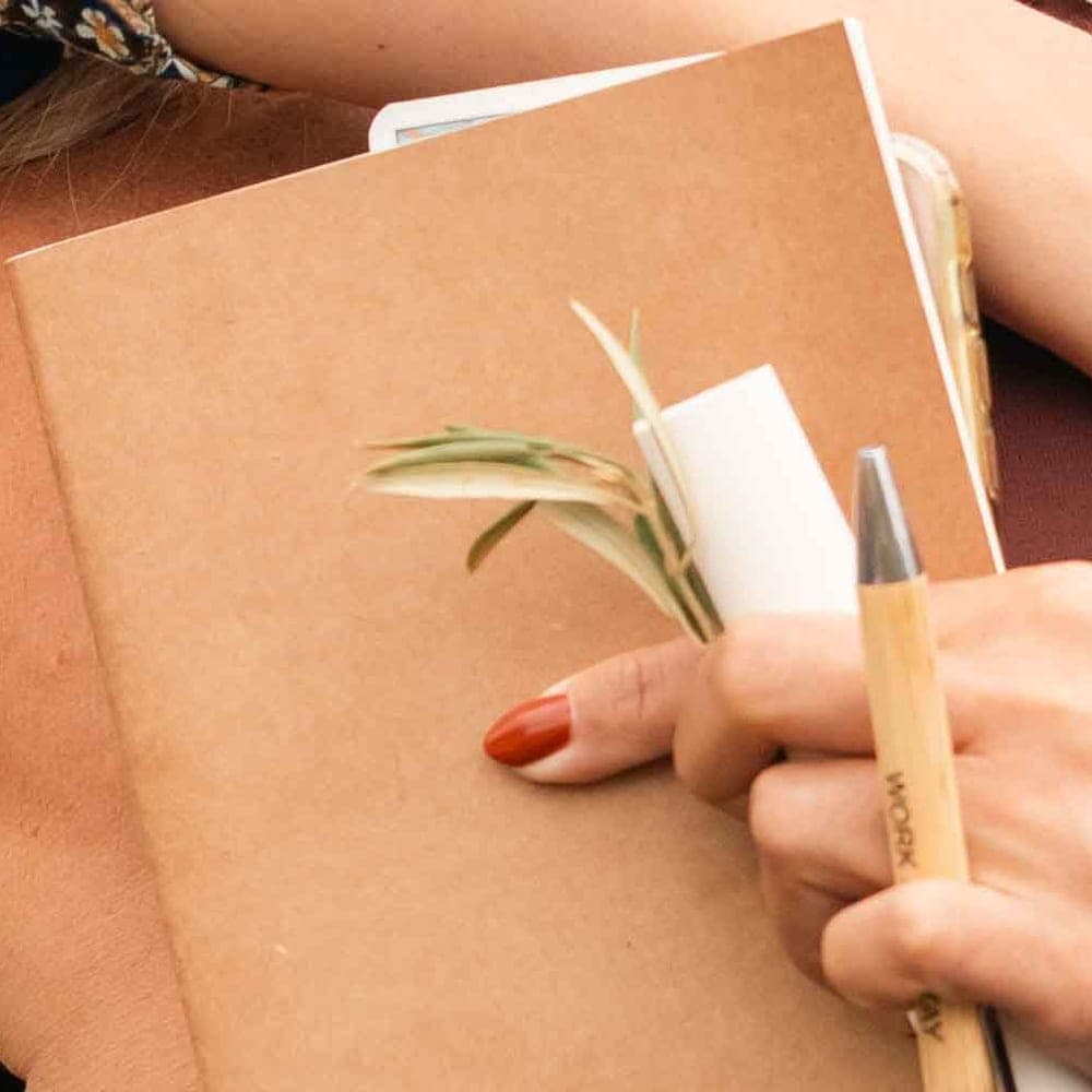 A kraft journal held against the body, a bamboo pen and a sprig of olive tucked into it