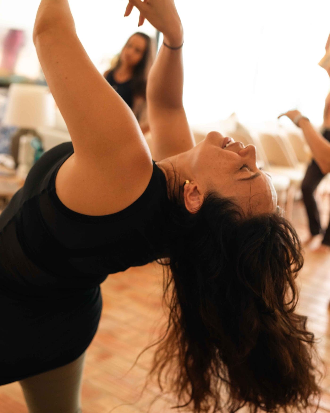 Camila Mello arching back with both arms raised, in bright window light